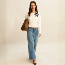 Load image into Gallery viewer, Woman wearing a white sweater with a black design, blue jeans, and holding a brown bag on a beige background