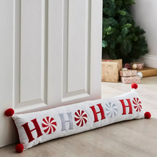 Load image into Gallery viewer, Decorative pillow with 'HO HO HO' text and red pom-poms on a wooden floor with a Christmas tree in the background.
