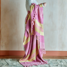 Load image into Gallery viewer, Person draped in a pink and yellow striped blanket against a neutral wall.