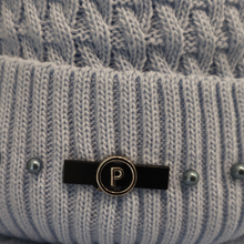 Load image into Gallery viewer, Close-up of a light blue cable-knit beanie with a black enamel and gold-tone metal pin featuring a letter 'P' in the center.
