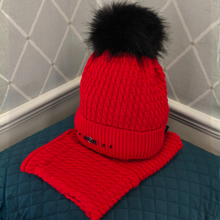 Load image into Gallery viewer, A bright red knit beanie hat with a large black faux-fur pom-pom and a matching red knit neck warmer, displayed on a quilted teal surface against a gray patterned wall.