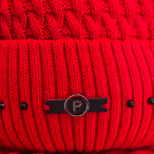 Load image into Gallery viewer, A close-up of a bright red ribbed knit beanie featuring a black metal bar with a circular silver-toned emblem containing the letter 'P', accented by small black beads on the side.