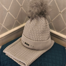 Load image into Gallery viewer, A light beige knit pom-pom beanie hat with a matching flat knit pom-pom band, displayed on a dark teal quilted cushion against a gray patterned wall.