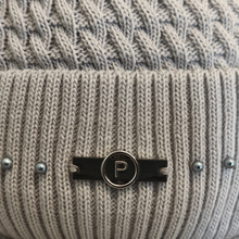 Load image into Gallery viewer, Close-up of a beige knitted beanie featuring a beautiful cable-knit pattern, finished with a decorative black enamel pin with gold-tone detailing and a central letter “P”.