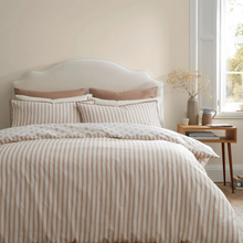 Load image into Gallery viewer, A neatly made bed in a softly lit bedroom, featuring a striped duvet cover and matching pillow shams in warm beige and cream tones, with coordinating blush pillows and a white upholstered headboard. A small wooden side table with a vase and books sits beside a bright window.
