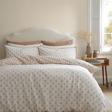 Load image into Gallery viewer, A neatly made bed in a softly lit bedroom, featuring a cream duvet cover and matching pillow shams with a small repeating leaf pattern, plus coordinating beige pillows. A white upholstered headboard, a wooden bedside table with books and a vase, and a window are visible.
