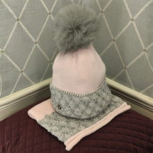 Load image into Gallery viewer, A light pink knit beanie hat with a large gray faux fur pom-pom, paired with a matching gray and pink knit neck warmer, displayed on a quilted burgundy surface against a gray patterned wall.