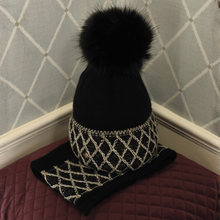Load image into Gallery viewer, A black knit beanie hat with a large black faux-fur pom-pom, featuring a decorative silver rhinestone diamond pattern band and a matching black-and-silver rhinestone scarf, displayed on a burgundy quilted surface against a patterned wallpaper background.
