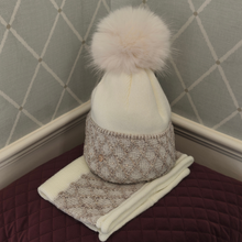Load image into Gallery viewer, A cream knit beanie hat with a large fluffy cream pom-pom, paired with a matching knit scarf, displayed on a quilted burgundy surface against a patterned gray wall.