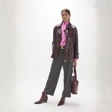 Load image into Gallery viewer, A model stands in a studio wearing a dark brown leather moto jacket over a bright pink high-neck blouse, paired with dark gray pinstripe wide-leg trousers, matching dark brown leather ankle boots, and holding a dark brown leather handbag.