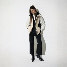 Load image into Gallery viewer, A model stands against a white wall wearing a beige and white puffer vest over a black top and black wide-leg trousers, paired with black pointed-toe boots.