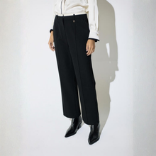 Load image into Gallery viewer, A model wearing black wide-leg cropped trousers, a white collared shirt, and black pointed-toe ankle boots, standing against a white wall with a strong shadow.