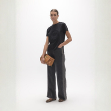 Load image into Gallery viewer, A model stands against a white studio background wearing a black graphic t-shirt, dark gray wide-leg jeans, leopard-print flat shoes, and holding a brown leather handbag.