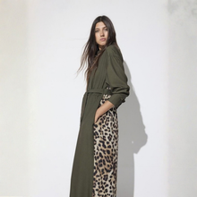 Load image into Gallery viewer, A woman modeling a long olive green belted coat over a leopard-print dress, standing against a plain white wall with a soft shadow.
