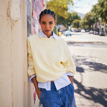 Load image into Gallery viewer, Woman wearing a light yellow sweater over a white shirt and blue jeans, leaning against a wall on a street.