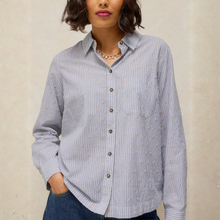 Load image into Gallery viewer, A woman wearing a light blue and white vertically striped button-up shirt with small embroidered details, paired with dark blue jeans, set against a neutral textured wall.
