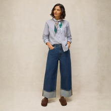 Load image into Gallery viewer, A woman stands against a textured beige studio backdrop wearing a light blue button-up shirt, wide-leg blue jeans with cuffed hems, brown loafers, and a patterned neck scarf.