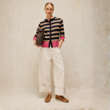 Load image into Gallery viewer, A model stands against a textured beige studio backdrop wearing a striped cardigan with navy, beige, and pink horizontal bands, paired with wide-leg cream trousers and brown suede boots. She holds a dark brown leather bag.