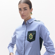 Load image into Gallery viewer, A woman wearing a light blue and white striped button-down shirt with a dark green plaid chest pocket featuring a gold crest logo. She is photographed against a clean, off-white studio background.