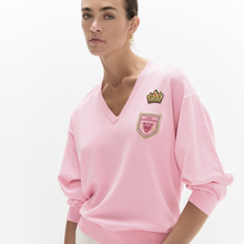 Load image into Gallery viewer, A woman wearing a light pink V-neck sweatshirt with embroidered school-style badges on the chest, photographed against a clean white studio background.
