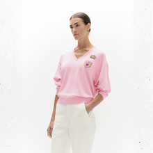 Load image into Gallery viewer, A woman wearing a light pink long-sleeve V-neck sweatshirt with embroidered patches on the chest, paired with white trousers, standing against a white textured wall.