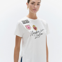 Load image into Gallery viewer, A woman wearing a white short-sleeve t-shirt with decorative embroidered patches and 'Made in Lofa' text on the front, set against a plain light gray background.