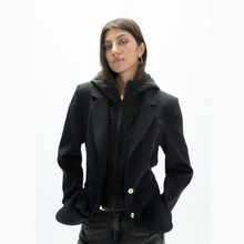 Load image into Gallery viewer, A woman wearing a black hooded jacket with structured shoulders and gold-tone buttons, layered over a black textured top and dark jeans, photographed against a plain white studio background.