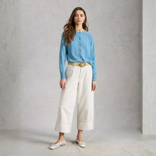 Load image into Gallery viewer, A woman stands in a studio wearing a light blue button-front cardigan, off-white cropped wide-leg pants, a tan belt, and white pointed flats against a textured light gray wall.