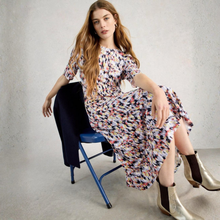 Load image into Gallery viewer, Woman in a colorful dress sitting on a blue chair against a plain background