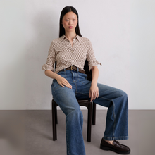 Load image into Gallery viewer, Woman sitting on a chair wearing a patterned shirt and blue jeans against a plain background