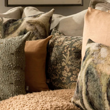 Load image into Gallery viewer, A collection of textured and patterned throw pillows arranged on a sofa in a cozy living room setting. The pillows feature a mix of solid camel, floral damask, and distressed linen patterns in earthy tones of beige, brown, and muted green.
