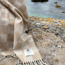 Load image into Gallery viewer, Beige checkered blanket with fringes on a rocky surface near water