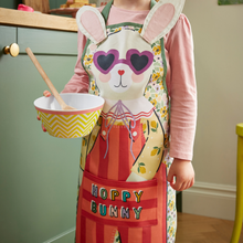 Load image into Gallery viewer, Child wearing a colorful 'Happy Bunny' apron holding a bowl and wooden spoon in a kitchen.