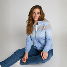 Load image into Gallery viewer, A woman sits cross-legged on a light gray studio floor wearing a light blue zip-up sweater with horizontal knit patterned stripes in white and taupe, paired with blue jeans.