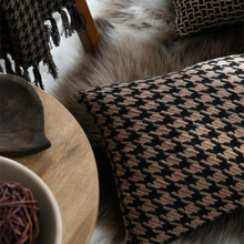 Load image into Gallery viewer, A square throw pillow with a black and beige houndstooth pattern, styled on a white faux fur rug next to a wooden bowl and a round wooden table.