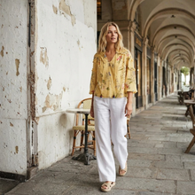 Load image into Gallery viewer, Woman in a yellow floral blouse and white pants standing in an outdoor setting with arches and tables.