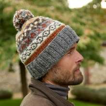 Load image into Gallery viewer, A man in profile wearing a knitted pom-pom beanie with a gray ribbed brim and a multicolor patterned upper section, photographed outdoors with blurred trees in the background.