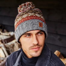 Load image into Gallery viewer, A man wearing a gray cable-knit beanie with a multicolor striped pom-pom and a small brown logo patch on the side, posed against a dark rustic wooden background.