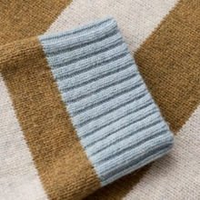 Load image into Gallery viewer, A close-up of a ribbed knit fabric cuff in a light dusty blue color, laid diagonally over a striped knit textile with brown and cream sections.