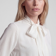 Load image into Gallery viewer, A close-up of a woman wearing a cream-colored blouse with a large bow tie-neck detail, set against a plain light gray background.