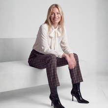 Load image into Gallery viewer, A woman sits on a white bench in a studio setting, wearing a cream long-sleeve blouse with a large bow at the collar, brown plaid cropped trousers, and black pointed-toe ankle boots.
