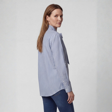Load image into Gallery viewer, A woman wearing a light blue and white vertically striped button-down shirt with a tie-neck detail, paired with dark blue jeans, photographed against a neutral light gray background.