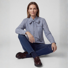 Load image into Gallery viewer, A woman sits cross-legged on a light gray studio floor wearing a blue and white vertically striped long-sleeve blouse with a large bow at the collar, dark blue jeans, and brown leather tassel loafers.
