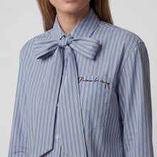Load image into Gallery viewer, A close-up of a model wearing a blue and white vertically striped button-down shirt with a large self-tie bow at the collar and embroidered script on the chest pocket.