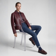 Load image into Gallery viewer, A model sits on a clear acrylic chair wearing a dark burgundy leather jacket, a patterned shirt, blue jeans, and brown leather loafers against a light gray studio background.