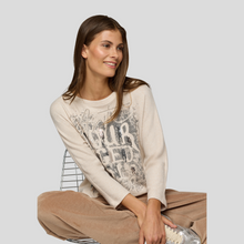 Load image into Gallery viewer, Woman wearing a beige sweater with text and brown pants sitting on a chair against a plain background