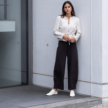 Load image into Gallery viewer, Woman in a white blouse and black pants standing against a gray wall.