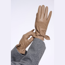 Load image into Gallery viewer, A pair of tan leather gloves with a bow detail at the wrist, worn on hands with a gray sweater visible, against a plain light background.
