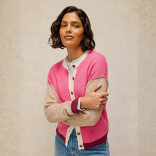 Load image into Gallery viewer, A woman wearing a color-block cardigan with a bright pink body, beige sleeves and collar trim, and a dark red ribbed hem and cuffs, paired with blue jeans against a textured beige background.