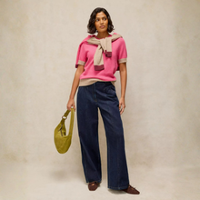 Load image into Gallery viewer, A woman stands against a warm beige studio backdrop wearing a bright pink short-sleeve knit top with beige and burgundy trim, dark wide-leg jeans, brown shoes, and carrying a muted olive green shoulder bag.